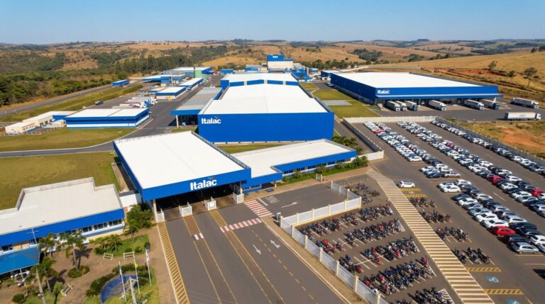 Vista aérea de uma fábrica industrial com galpões e pátio amplo, estacionamentos com muitos veículos e caminhões, cercada por áreas rurais ao fundo.